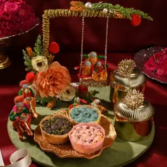 This whimsical Teej hamper features a miniature swing (jhoola), colourful ghewars, and ornate golden jars. It’s a playful and traditional gift that captures the essence of the festival.