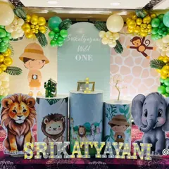 A safari-themed "Wild One" party featuring a little explorer character and friendly jungle animals. The setup includes an arched backdrop, custom-printed plinths, and a full balloon garland in green and gold.