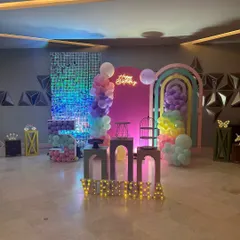 A modern and chic rainbow themed birthday party setup. The decor includes a pink backdrop, a shimmer wall, and artistic rainbow-shaped arches with pastel balloon accents.
