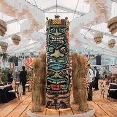 A massive, multi-faced Tiki totem pole stands as the central feature of the event space. Surrounded by pampas grass, this impressive sculpture is a key element of the authentic tropical atmosphere.