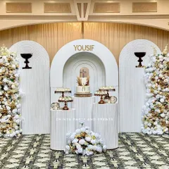 An elegant and serene stage for a First Holy Communion. This setup uses white 3D ridged panels, tall pedestals for the cake and offerings, and lush white and gold floral arrangements for a sophisticated look.