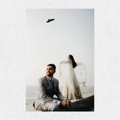 A creative shot from a beachside pre-wedding session. The groom sits in the foreground while the bride stands behind him with her dupatta flying in the wind, like a scene from a movie.