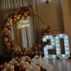 An elegant 20th birthday setup with a circular balloon arch in white and rose gold. A "Happy Birthday" neon sign and large marquee numbers add a warm glow to the celebration.