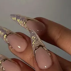 A close-up of extremely long, clear stiletto extensions. The tips are adorned with a 3D gold textured design, creating a look that is both delicate and fierce.