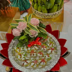 A colorful platter of assorted Indian sweets. The arrangement is placed on a decorative lotus base and topped with pink roses, making it a beautiful and delicious gift for any celebration.