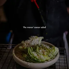 A chef drizzles dressing over the Manor Caesar salad. This action shot highlights the freshness of the ingredients and the care that goes into preparing even the simplest dishes.