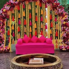 A stunning Mehendi seating arrangement with a vibrant pink sofa set against a backdrop of hanging yellow and orange tassels and a lush floral arch. The traditional carved fire pit adds an authentic touch to the festive decor.