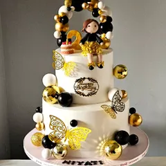 A chic and glamorous cake in a black, gold, and white theme for a second birthday. The design features a balloon garland, shimmering butterflies, and a custom-made girl figurine in a sparkling dress.
