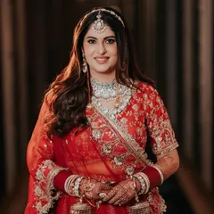 A classic, forward-facing bridal portrait. The soft lighting highlights her intricate jewelry and the rich color of her red lehenga, capturing her calm and radiant beauty just before the ceremony.