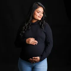 A simple and beautiful portrait of a woman in a black top and jeans, cradling her bump against a solid black background. Her gentle smile and the classic styling create a warm and timeless feel.