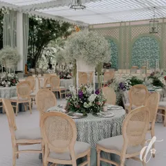 A beautifully set table featuring a tall, airy centerpiece of baby's breath and a lush base of mixed florals. The classic wooden cane-back chairs and patterned tablecloth complete this picture of garden party elegance.