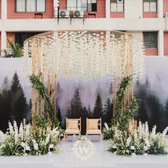 A modern minimalist mandap that perfectly blends nature and contemporary design. The structure features a backdrop of a misty forest, a canopy of hanging white florals, and lush green arrangements.