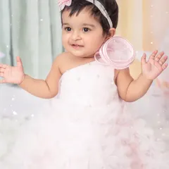 A baby girl in a beautiful white dress, surrounded by a dreamy pastel rainbow and cloud-themed set for her first birthday.