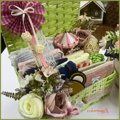 A custom baby hamper designed as a keepsake. This vibrant green woven basket is filled with a variety of textures and items, including a miniature carousel, folded baby clothes, and decorative floral elements.