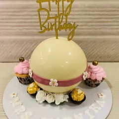 A surprise inside! This white chocolate piñata sphere cake is designed to be smashed open, revealing a fun treat. It's surrounded by matching cupcakes and Ferrero Rocher.