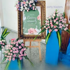 A personalized welcome sign for a naming ceremony, framed in ornate gold and topped with a lush arrangement of pink and purple flowers. The surrounding floral stands continue the color theme, creating a cohesive and inviting entrance.