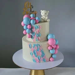 A classic baby shower or gender reveal cake in pink and blue. This two-tiered design features a cascade of balloons, a textured buttercream effect, and a sweet teddy bear topper.
