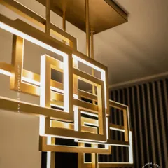 A multi-layered chandelier composed of intersecting rectangular frames with a brushed gold finish. The integrated LEDs cast a warm, ambient light, creating a dynamic and architectural statement piece for modern interiors.