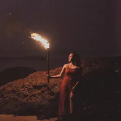A full-length portrait of the bride-to-be in a red dress, standing by a rock and holding a flaming torch. The warm light of the flame illuminates her against the dark beach, creating a bold and beautiful image.