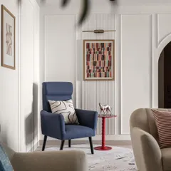 A cozy reading corner in the living room, featuring a high-back armchair in a striking blue. The wall paneling adds subtle texture, while the abstract art and small red side table provide a playful touch of color.