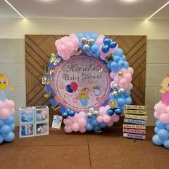 A complete baby shower setup for 'Hiral's Baby Shower'. The decor features a circular backdrop with a pink and blue balloon arch, baby-themed cutouts, and 'BABY' boxes.