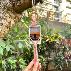 A single pink macrame polaroid holder hanging from a tree, showcasing a beautiful sunset photo. It's a perfect, personalized gift to remember a special trip or moment.