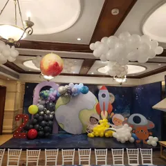 An incredible outer space themed birthday party. The stage is set with a galaxy backdrop, planet cutouts, a rocket ship, astronauts, and balloon clouds to create a cosmic adventure.