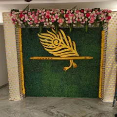 This backdrop features a golden Krishna flute and peacock feather cutout against a green grass wall, topped with a dense arrangement of pink and white flowers.