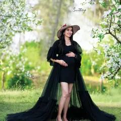 A chic and stylish outdoor portrait. The mother-to-be, in a black dress and hat, stands amidst blooming white flowers, creating a beautiful contrast and a high-fashion look.