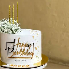 A simply aesthetic birthday cake. The clean white base is decorated with fresh baby's breath, gold leaf, and a modern acrylic "Happy Birthday" topper.