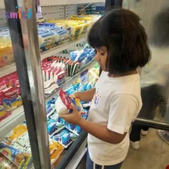 A child examines a packet of paneer in the refrigerated section, learning about different types of food and where they are stored in a supermarket.