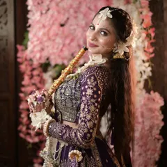 Radhe Radhe. A beautiful Janmashtami look featuring a purple lehenga, fresh flower jewellery, and delicate face art. The model holds a flute, completing the divine portrayal.