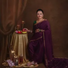 A classic portrait of a mother-to-be in a deep purple saree, styled in a traditional way. She stands next to a table with fruits and brass lamps, evoking a sense of timeless grace.