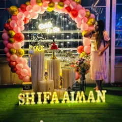 A beautiful 'Bride to Be' party setup on a terrace. The decoration includes a pink and gold balloon arch, a neon sign, and a full display of props on an artificial grass surface.