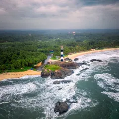 The iconic Kaup lighthouse standing defiant against the powerful monsoon waves. This aerial shot captures the raw energy of the Arabian Sea and the solitude of the lighthouse, a beacon on the rugged coast.
