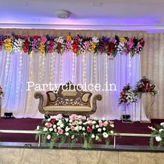 This is a full stage view for a Half-Saree function or similar puberty ceremony. The colorful floral header and simple white backdrop provide a beautiful setting for the young lady being celebrated.