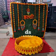 This is another one of our popular Haldi setups, arranged outdoors. It features an artificial grass backdrop with a red 'हळद' sign, marigold and tuberose hangings, and a large decorative Urli for the ceremony, all available for your event.