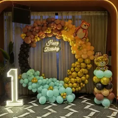 A cute and trendy teddy bear themed first birthday. The circular balloon arch uses a modern color palette of brown, gold, and teal, with a 'Happy Birthday' neon sign and a light-up number '1'.