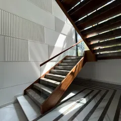 The staircase at Gulmohar Farmhouse is a study in light and texture. Sunlight streams through the large window, casting shadows across the stone steps and the adjacent wall, which features a rhythmic pattern of smooth and fluted panels.
