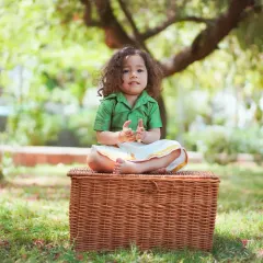 A full-body shot of a toddler in a traditional South Indian outfit, sitting on a wicker basket during an outdoor shoot. The natural setting is perfect for cultural portraits.