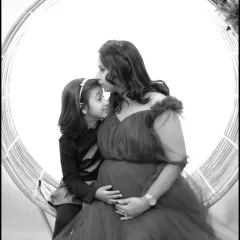 A tender black and white moment between a mother and her daughter, seated in a circular prop. The mother kisses her daughter's forehead, celebrating their bond.