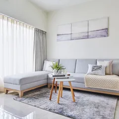 The living room is a tranquil sanctuary with crisp white walls and a sophisticated light grey sofa. A wooden coffee table and sheer curtains that filter sunlight complete this serene and blissful haven.