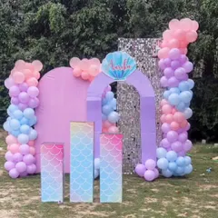 An outdoor mermaid theme setup perfect for a daytime party. It features a pink arch backdrop against a silver sequin wall, with balloon columns in pastel shades and pedestals with a mermaid scale pattern.