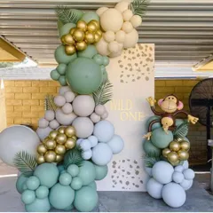 A "Wild One" first birthday setup featuring a stylish organic balloon garland in sage green, white, and gold, accented with tropical leaves and a playful monkey.