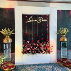 A "Love in Bloom" neon sign set within a white frame and surrounded by flowers, creating a modern and romantic photo booth for the couple and guests.