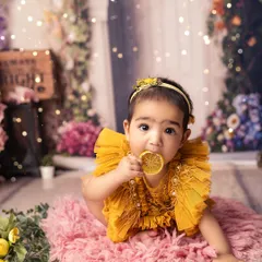 A little sunshine tasting a slice of sweetness. The combination of the floral background, her bright yellow dress, and the lemon prop creates a vibrant and joyful image.