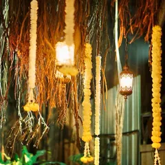 An atmospheric shot of rustic decor at night. Garlands of marigolds and tuberoses hang from banyan tree roots, interspersed with glowing antique lanterns that create a warm and enchanting mood.