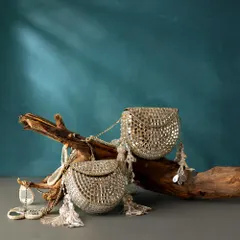 A duo of silver clutches, heavily embellished with mirror work and finished with delicate tassels. They are displayed on a piece of driftwood, highlighting their artistic quality.