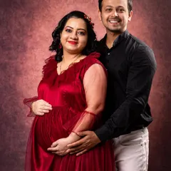 A classic studio maternity portrait against a textured backdrop. The mother-to-be is radiant in a red gown, creating a timeless and elegant image celebrating her pregnancy.