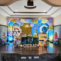 For this 5th birthday, I created an elaborate Little Krishna theme on a full stage. The decoration features two large printed backdrops, a central gold shimmer panel, a multi-colored balloon arch, character statues, and illuminated marquee letters for the child's name.
